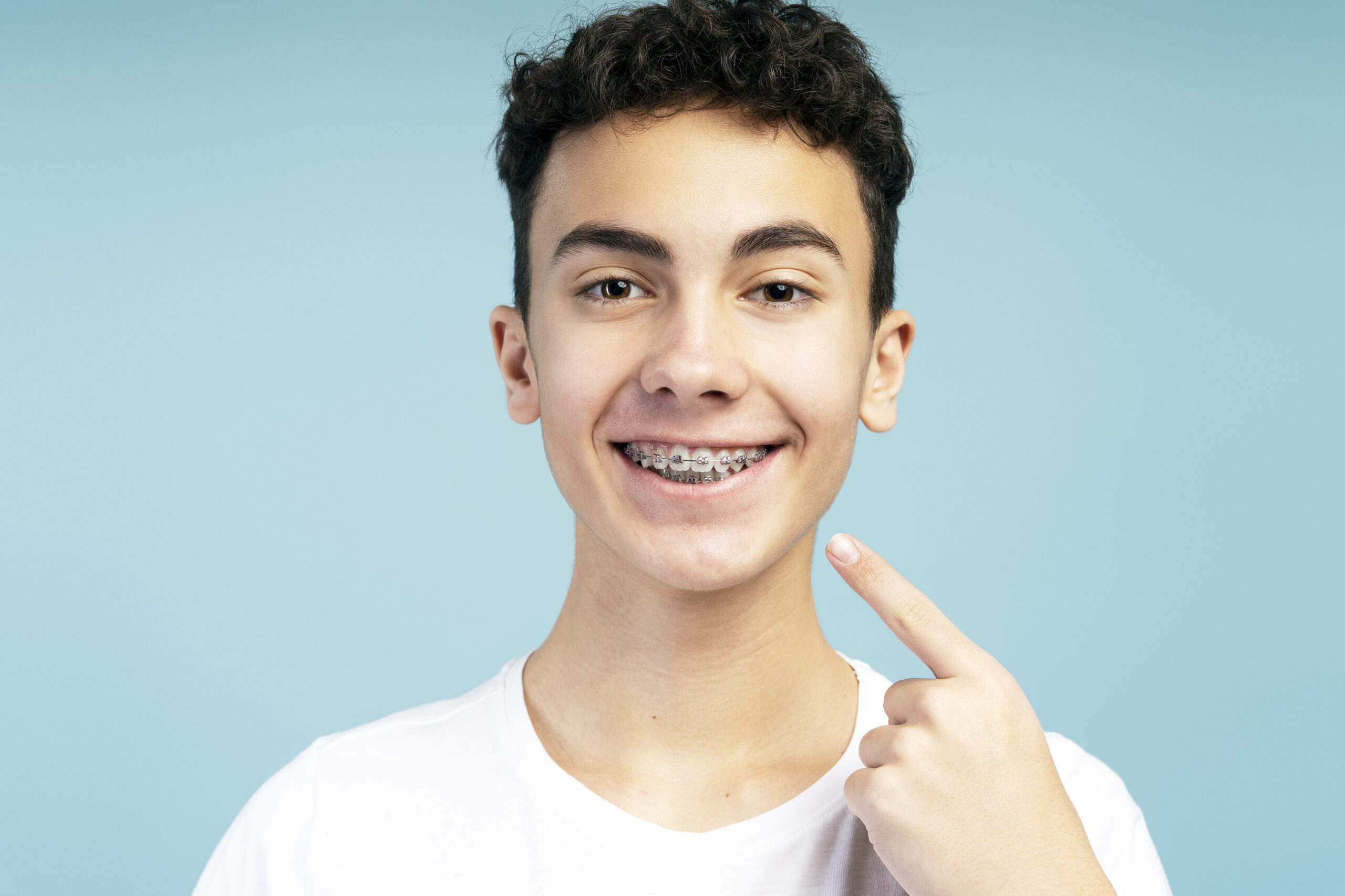 Positive attractive boy with braces pointing finger to mouth, looking at camera. Handsome teenager posing isolated on blue background. Concept of dental, health care